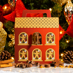 A red gingerbread house shaped gift box with a handle, decorated with gold window frames and gingerbread men illustrations, set on a snowy surface with cinnamon sticks and chocolate pieces. Christmas trees, ornaments, a gold gift box, and red ribbons are visible in the background.