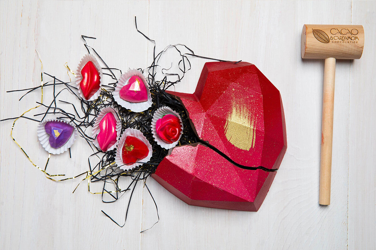 A heart-shaped smash cake with a dark chocolate base, decorated with red and gold accents, and filled with small chocolate pieces. A wooden mallet is also present, positioned to the right of the cake.