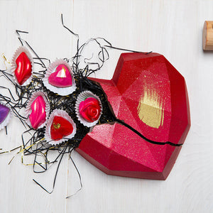 A heart-shaped smash cake with a dark chocolate base, decorated with red and gold accents, and filled with small chocolate pieces. A wooden mallet is also present, positioned to the right of the cake.