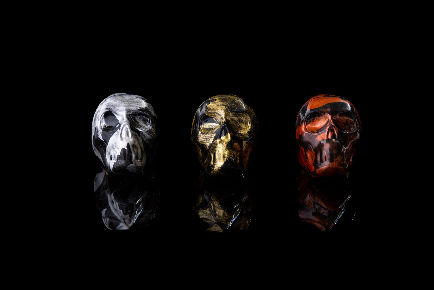 Three different colored caramels designed to represent skulls, placed on a dark background. From left to right: copper skull with raspberry caramel, gold skull with salted caramel, and silver skull with dark chocolate caramel.