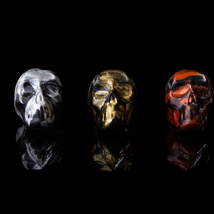 Three different colored caramels designed to represent skulls, placed on a dark background. From left to right: copper skull with raspberry caramel, gold skull with salted caramel, and silver skull with dark chocolate caramel.